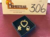 Three gold pendants in a black box on burgundy cloth: heart pendant, loop with pearl, double letter B.