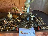 Photo showing entire lot on table with multiple brass and metal decorative items including figurines, candleholders, fan, medallions and hardware