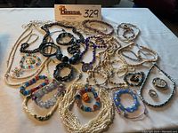 Photo showing assorted necklaces, bracelets, rings, pins including beads, pearls, shells, colorful and black beads laid out on white surface.