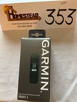 Front view of boxed Garmin Vivofit 4 in black with visible digital time display on packaging, showing branding and size S-M, with note about 1-year battery life.