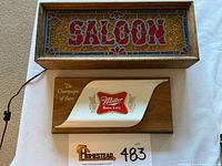 Photo showing two vintage signs on white surface: a Saloon sign with wooden frame and stained glass style design above, and a Miller High Life sign on wood panel below.
