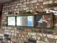 Full frontal view of vintage three-panel wall mirror mounted on brick wall, showing ornate gold wood frame and reflections in mirror panels.