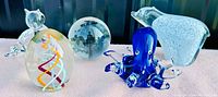 Collection of six art glass paperweights and figurals on shelf, showing various shapes and colors including blue octopus, clear cat, abstract shapes, and colorful oval paperweight.