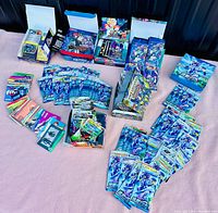 Wide view showing multiple booster packs, cards, and boxes from Pokemon, Dragon Ball Z, Yu-Gi-Oh!, Fortnite organized on pink surface.