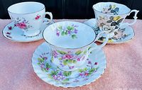 Front view of three fine bone china tea cups with matching saucers displaying floral patterns in pink, purple, and beige tones with gold trim.