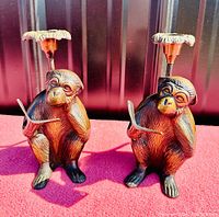 Front view of two bronze monkey candle holders on red surface with one holding a book in each hand and floral candle holder above.