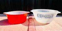 Photo showing the two bowls side by side: red Pyrex casserole dish and white Glasbake milk glass Currier & Ives bowl on a pink cloth.
