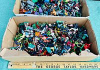 Two cardboard trays filled with assorted small plastic toy figurines on greenish surface with ruler in front