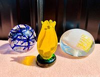 Three vintage art glass paperweights arranged side by side on a pink surface, showing a clear round orb with blue swirls, a yellow tulip-shaped paperweight, and an oval clear glass paperweight with mosaic patterns.