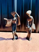 Photo showing two vintage cast iron Geisha girls sculptures with geometric styling, black finish, standing on a textured surface with sunlight highlighting details.