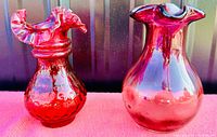 Two vintage cranberry glass vases side by side on a red surface against a dark background, showing the ruffled tops and overall shapes.