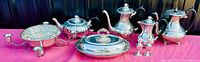 Wide view showing all silver plated items on a magenta cloth, including teapots, coffee pot, milk jug, salt and pepper shakers, oval covered serving tray, centerpiece bowl with flower frog, and warming tray with candle holders.