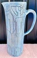 Front view of pottery jug showing abstract human figures etched on blue/green glaze surface.