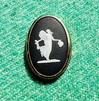 Close-up view of the vintage Wedgwood black jasperware porcelain cameo brooch pendant laid on green fabric, showing white classical figure on black background.