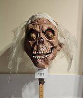Front view of Cryptkeeper rubber Halloween mask on stand with measuring ruler underneath showing approximately 11 inches in height. Mask has detailed skeletal facial features and synthetic white hair.