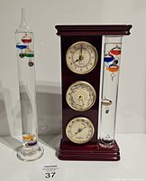 Photo showing both Galileo thermometers: one free-standing glass thermometer and one wooden framed weather station with clock, barometer, and hygrometer.