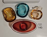 Photo showing all four colorful Murano-style art glass trays: deep red elongated oval, amber apple shape, amber kidney shape, and round blue tray