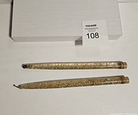 Two lucite candles with gold flecks inside, placed horizontally on a white surface with an auction number card displayed behind them.