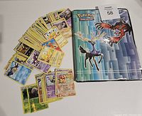 A collection of Pokémon trading cards next to a binder featuring Xerneas and Yveltal on the cover.