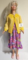 Front view of very tall Barbie-style doll wearing yellow knit sweater and floral dress with pink shoes.