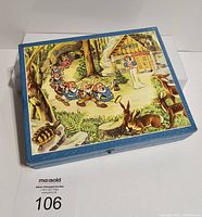 Closed puzzle box with blue border and image of dwarfs approaching Snow White's cottage with woodland animals in foreground. Puzzle box is square shaped with clasp.