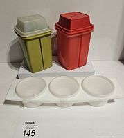 Two rectangular Tupperware containers (green and red) and a white tray with three round condiment containers with lids arranged in front of a white background.