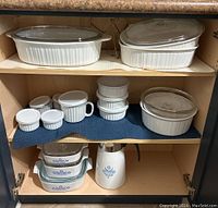 Shelf with large oval and round white ribbed French White bakeware dishes with glass lids on top shelf; mid shelf holds small ramekins, mug, and bowls; bottom shelf contains Corningware Cornflower blue patterned casseroles and coffee pot.