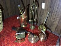 Brass fireplace tool stand with shovel, brush, poker, tongs, copper pitcher with ceramic handles, copper pot with lid and two kettles, antique fire bellow, and a small iron stove-shaped piece on red surface by wood panel wall.