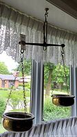 Brass hanging balance scale with two bowls suspended by chains in front of window with lace curtains.