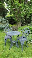 Front view of the bistro set showing two matching cast iron chairs and round table placed on grass in a garden with trees in the background.