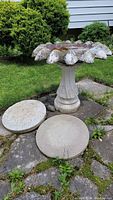 Full view of weathered concrete birdbath and two round concrete patio stones placed on the ground with stone pavers and grass surroundings.