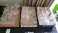 Overview of three trays containing multiple clear glass stemware and barware of various styles with detailed cut glass patterns arranged inside.
