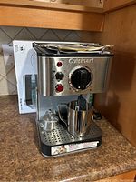 Front view of the stainless steel Cuisinart espresso maker on countertop showing control knob, two red indicator lights, frothing pitcher and tamper on drip tray. Box and instruction manual are placed behind the machine.