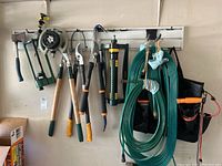 Wide shot of garden tools hanging on a wall rack including hand pruners, loppers, sprinkler, hose, and storage bag