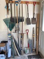 Wide view showing multiple long-handled gardening tools hanging on wall including shovels, rakes, pitchfork, saw, bamboo stakes and rolls of wire and fabric on floor.