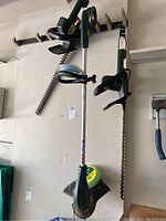 Two green Black & Decker hedge trimmers with long blades hanging on wall, Yardworks weed eater with curved guard below them, spool of trimmer line beneath weed eater.