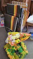 Two folded vintage mesh lawn chairs with brown mesh seats and backrests featuring yellow and orange stripes. Two colorful retro outdoor cushions with floral patterns placed in front.