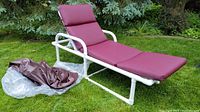 Full view of lounge chair with maroon cushion on frame and plastic sleeve holding additional cushion parts on grass.