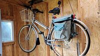 Side view of silver Raleigh sports bike hanging by pulley system inside wooden shed. Visible Shimano drivetrain, full metal fenders, front wire basket, and rear pannier bags.