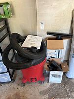 Wide view of red Craftsman wet/dry shop vacuum with large black hose coiled on top. Various vacuum attachments in cardboard boxes beside it.