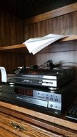 Sony CD changer player model CDP-CE500 with remote and Optimus LAB-1100 turntable stacked above it on wooden shelving. RCA cables visible on top of turntable.