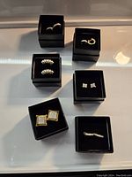 Six pairs of pierced earrings displayed individually in black boxes, showing different styles, sizes, and metal finishes with clear stones.