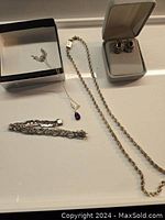 Photo shows the full lot of vintage sterling silver jewelry: earrings in a presentation box, a rope chain necklace, a rope bracelet, and a delicate necklace with purple pendant inside a black box.