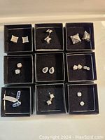 Nine pairs of assorted silver-toned stud earrings with various clear and blue stones placed in nine black boxes, arranged in a three by three grid.