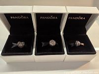 Three Pandora charms in their original white boxes with black velvet interiors. Left charm is heart-shaped with floral cutouts. Center charm is round with clear crystals. Right charm is round bead with alternating black and clear stones.