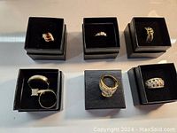 Photo of seven gold-tone rings in black boxes arranged on a surface showing various designs and stones.