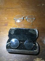 Top view of two pairs of antique eyeglasses, one resting on an old metal and velvet-lined case, the other pair octagonal shaped metal wireframe glasses on wooden surface.