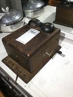Side and top angle view of the solid oak phone ringer box showing two black metal bells on top, the crank handle on the side, and wiring at the base.