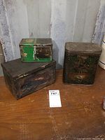 Three antique tins displayed on wooden surface showing aged wear and scuffs.