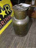 Solid brass canister standing on floor next to a display sign, showing its full height and shape with the lid on.